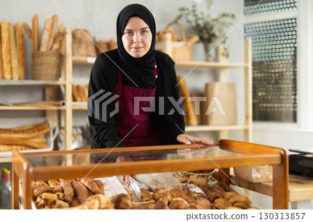 Muslim girl seller employee puts croissants in window, arranges display of goods at bakery. 130313857
