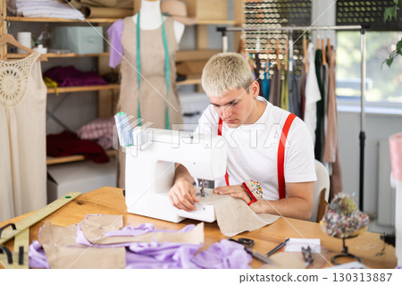 Attentive man tailor sewing textile using sewing machine sitting at workplace in fashionable sewing atelier 130313887