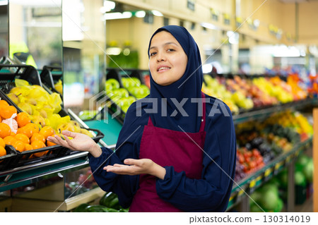 Female seller in hijab invites you to go to showcases with fresh fruits Female seller in hijab invites you to go to showcases with fresh fruits 130314019