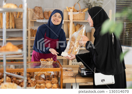 Female bakery salesperson in traditional Islamic dress serves bag of baguettes and bread to a girl in niqab 130314070