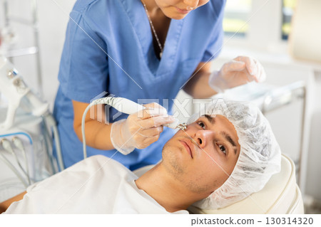 Young female cosmetologist doing microcurrent procedure to young man 130314320