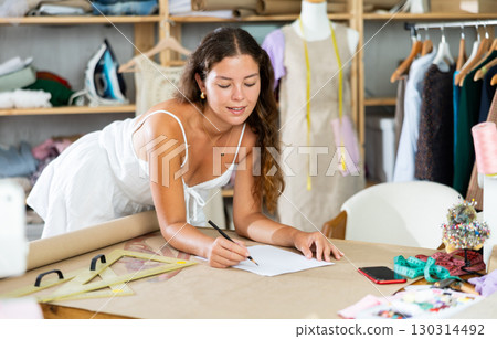 Dreamy seamstress drawing sketches leaning over table in studio Dreamy seamstress drawing sketches leaning over table in studio 130314492