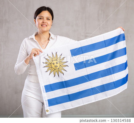Reliant positive young woman holds big national flag of Uruguay with confident smile showing teeth Reliant positive young woman holds big national flag of Uruguay with confident smile showing teeth 130314588