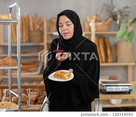 Female shopper in muslim clothing buys delicious pastries and checks the calorie count using smartphone 130314593