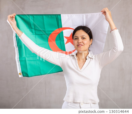 Reliant positive young woman holds big national flag of Algeria with confident smile showing teeth. Reliant positive young woman holds big national flag of Algeria with confident smile showing teeth. 130314644