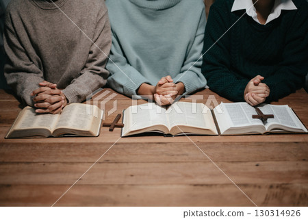 Group of Christians sit together and pray around a wooden table with blurred open Bible pages in their homeroom. 130314926