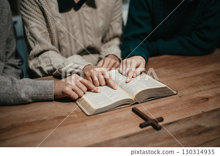 Group of Christians sit together and pray around a wooden table with blurred open Bible pages in their homeroom. 130314935
