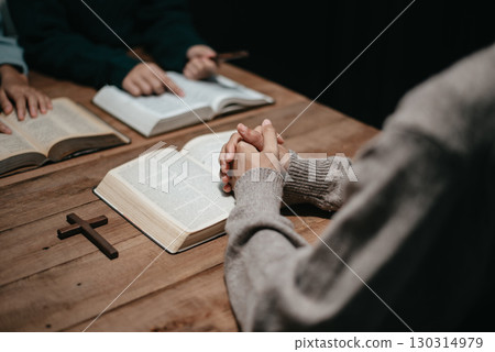 Group of Christians sit together and pray around a wooden table with blurred open Bible pages in their homeroom. 130314979