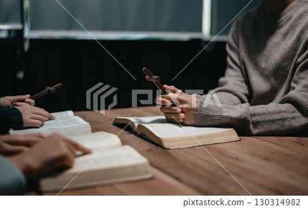 Group of Christians sit together and pray around a wooden table with blurred open Bible pages in their homeroom. 130314982