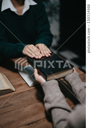 Group of Christians sit together and pray around a wooden table with blurred open Bible pages in their homeroom. 130314986