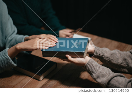 Group of Christians sit together and pray around a wooden table with blurred open Bible pages in their homeroom. 130314990