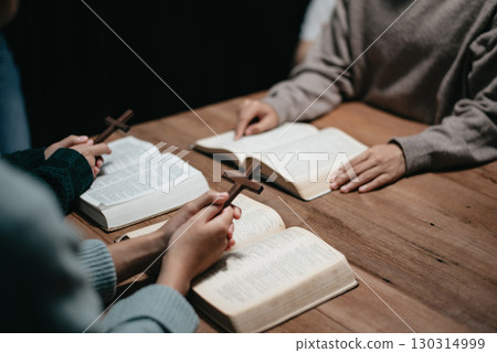 Group of Christians sit together and pray around a wooden table with blurred open Bible pages in their homeroom. Group of Christians sit together and pray around a wooden table with blurred open Bible pages in their homeroom. 130314999