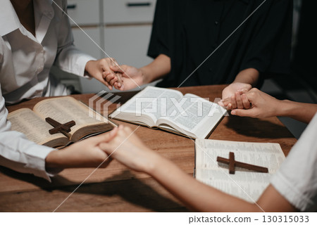 Group of Christians sit together and pray around a wooden table with blurred open Bible pages in their homeroom. 130315033