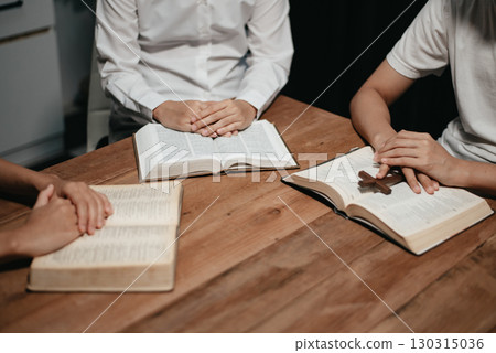 Group of Christians sit together and pray around a wooden table with blurred open Bible pages in their homeroom. 130315036