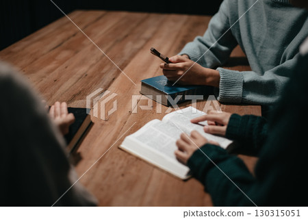 Group of Christians sit together and pray around a wooden table with blurred open Bible pages in their homeroom. 130315051