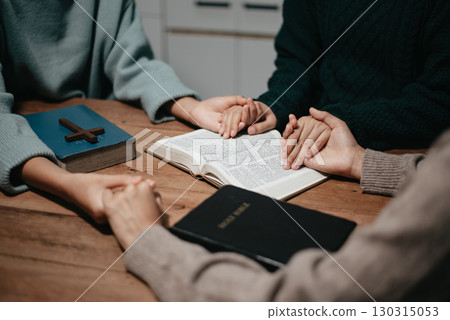 Group of Christians sit together and pray around a wooden table with blurred open Bible pages in their homeroom. Group of Christians sit together and pray around a wooden table with blurred open Bible pages in their homeroom. 130315053