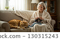 Senior woman knitting at home in a cozy living room with natural light and warm blanket, cat sleeping nearby. 130315243