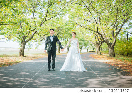 一對穿著禮服的情侶手牽手沿著公園的綠樹成蔭的大道散步（婚禮、婚紗照、新娘和新郎） 130315807