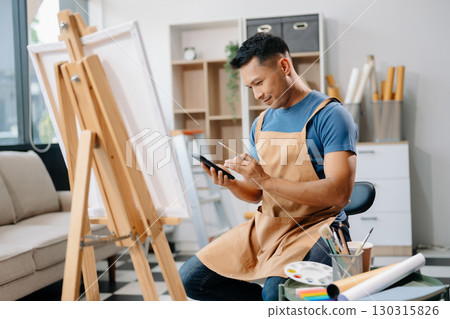 Asian male painter do artwork in art workshop, painting supplies, oil pastels, two canvas easel, creative space with paintbrush 130315826