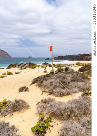 A scenic beach scene at La Graciosa, Lanzarote, with a red flag signaling potential hazards near the shore and a life ring ready. A scenic beach scene at La Graciosa, Lanzarote, with a red flag signaling potential hazards near the shore and a life ring ready. 130319340