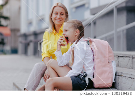 Young student eats apple with backpack and mother nearby before class, showing healthy habits on back to school morning. Young student eats apple with backpack and mother nearby before class, showing healthy habits on back to school morning. 130319428