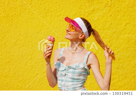 Joyful woman with ice cream cone in sunglasses and visor smiling on hot summer day against vibrant yellow wall Joyful woman with ice cream cone in sunglasses and visor smiling on hot summer day against vibrant yellow wall 130319451
