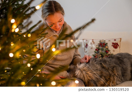 Woman decorating Christmas tree with lights while dog resting on sofa indoors Woman decorating Christmas tree with lights while dog resting on sofa indoors 130319520