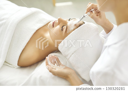 Female cosmetologist applying moisturizing mask with brush to a young woman in spa salon. Female cosmetologist applying moisturizing mask with brush to a young woman in spa salon. 130320281