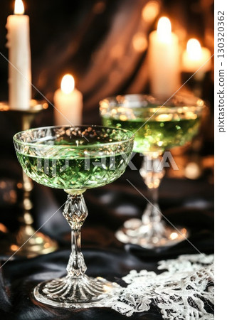 Glowing emerald cocktails in crystal coupes nestled against candlelight, casting eerie shadows on black satin for haunting halloween ambiance 130320362