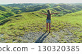Young woman tourist embracing freedom on Sumba Island, Indonesia, with arms outstretched in an idyllic landscape of lush green hills, basking in the warm glow of sunset 130320385