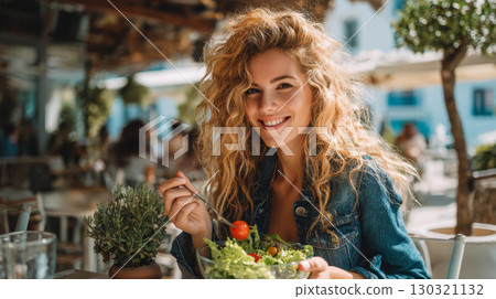 Young woman with curly hair smiling while eating fresh salad with cherry tomato at outdoor cafe on sunny day 130321132