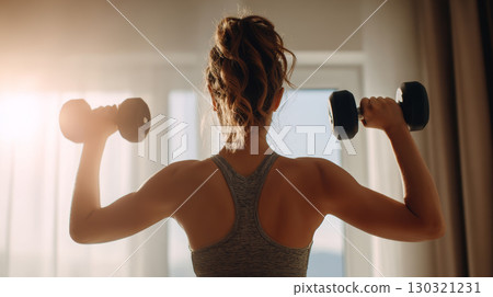 Young woman lifting dumbbell weights in bright room with sunlight, showing strong back muscles and fitness motivation 130321231