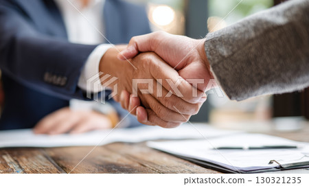Two business people shaking hands over wooden table with documents, expressing agreement and cooperation in professional setting Two business people shaking hands over wooden table with documents, expressing agreement and cooperation in professional setting 130321235