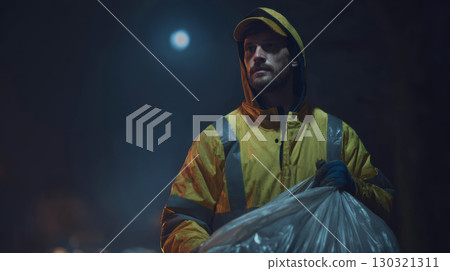 Man in yellow raincoat holding garbage bag at night with full moon in background, showing determination and focus 130321311