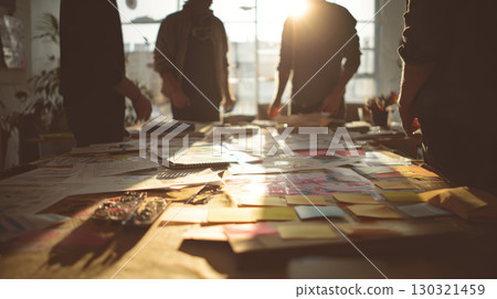 Brainstorming session with people standing around table covered with papers and sticky notes in warm sunlight 130321459