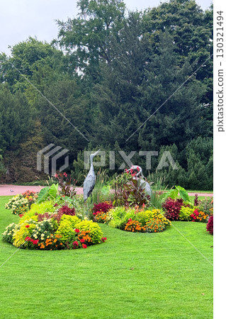 A landscaped city park features vibrant flower beds and a bird sculpture surrounded by lush greenery. Urban biodiversity, green infrastructure, ecological landscaping, pollinator habitats 130321494
