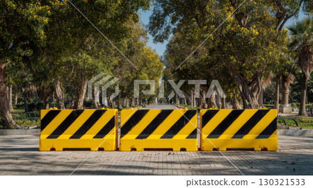 Temporary construction barricade with black and yellow stripes blocking tree lined pathway in sunny park, creating safe and controlled area 130321533