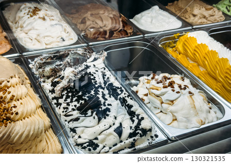 Multiple gelato flavors in a metal display case including cookies and cream, pistachio, and caramel swirl. Artisanal desserts, flavor diversity, indulgent treats, local gelateria culture 130321535