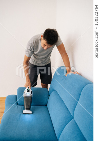 Vertical portrait of homeowner male using handheld vacuum cleaner to remove dust and dirt from blue sofa, emphasizing importance of cleanliness and hygiene in home maintenance. 130322764