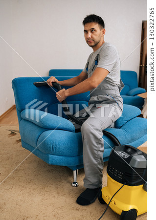 Full length vertical portrait of professional cleaning service employee wearing uniform using vacuum cleaner to clean blue sofa in living room, ensuring spotless and dust-free environment. Full length vertical portrait of professional cleaning service employee wearing uniform using vacuum cleaner to clean blue sofa in living room, ensuring spotless and dust-free environment. 130322765