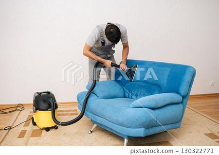 Wide shot of professional cleaning service worker removing dirt and stains from blue upholstered sofa using extraction washing vacuum cleaner in living room. Concept of domestic work, housekeeping. 130322771