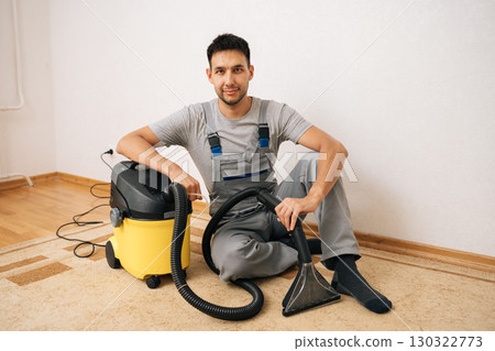 Portrait of professional cleaning service employee in overall sitting on floor next to washing vacuum cleaner, ready for thorough residential floor cleaning and sanitizing service, looking at camera 130322773