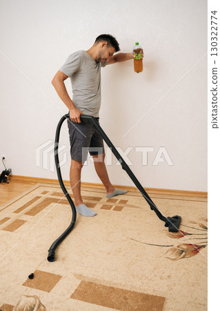 Tired young man vacuuming messy carpet while holding beer bottle and leaning against wall, feeling aftermath of lively party at home, showcasing humorous take on domestic chores. 130322774