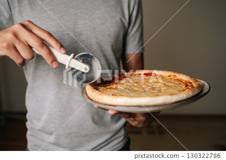 Close-up cropped shot of unrecognizable man wearing gray t-shirt holding round pizza on plate, cutting pizza using cutter wheel tool, ready to serve delicious slices for sharing at home, closeup. Close-up cropped shot of unrecognizable man wearing gray t-shirt holding round pizza on plate, cutting pizza using cutter wheel tool, ready to serve delicious slices for sharing at home, closeup. 130322786