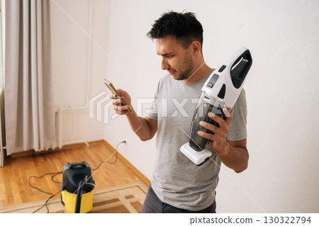 Portrait of focused male janitor wearing gray t-shirt and shorts, standing on carpet, using smartphone and holding cordless handheld vacuum cleaner, preparing to clean house. Portrait of focused male janitor wearing gray t-shirt and shorts, standing on carpet, using smartphone and holding cordless handheld vacuum cleaner, preparing to clean house. 130322794
