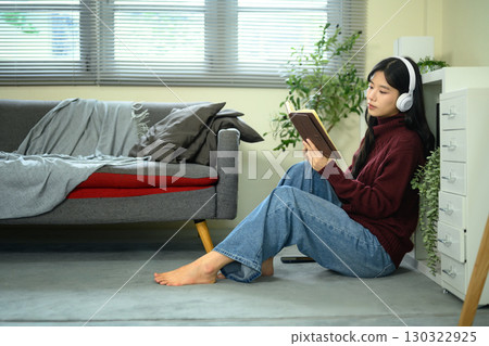 Relaxed Asian woman reading a book while listening to music, sitting comfortably on the floor 130322925