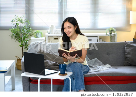 Smiling woman taking notes while attending an online meeting with laptop in a living room 130322937