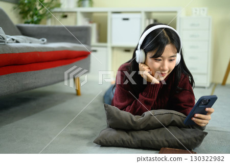 Young woman lying on the floor at home, wearing headphones and using her smartphone 130322982