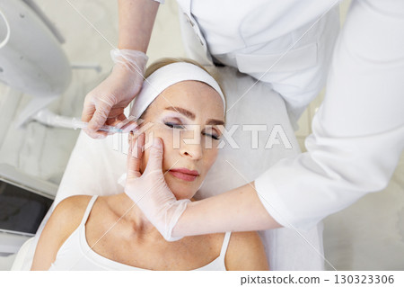 Senior woman receiving aesthetic facial injection by professional cosmetologist in white coat and gloves during anti-aging treatment procedure in modern clinic or beauty salon, lying on table 130323306