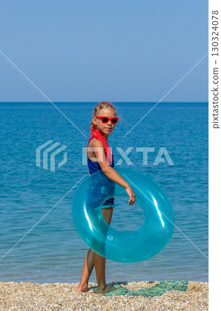 Girl with red braids and red sunglasses standing with blue inflatable ring on pebble beach, looking at the camera Girl with red braids and red sunglasses standing with blue inflatable ring on pebble beach, looking at the camera 130324078